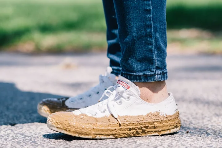 Close-up of white fabric sneakers with mud on the soles, standing on a sunlit pavement