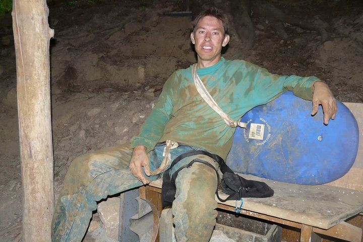 Person covered in mud sitting on a bench indoors, wearing a green shirt and muddy pants, with a blue helmet nearby.