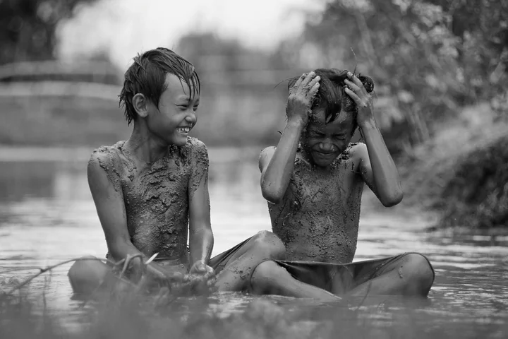 Two children covered in mud sit in shallow water outdoors, smiling and playing as mud coats their skin.