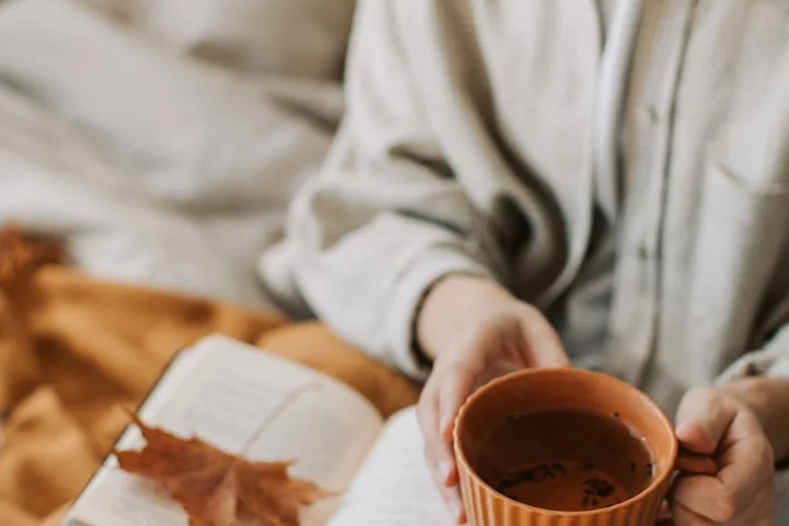 Close-up of a person holding an orange ceramic mug with tea, beside an open book on a soft blanket.