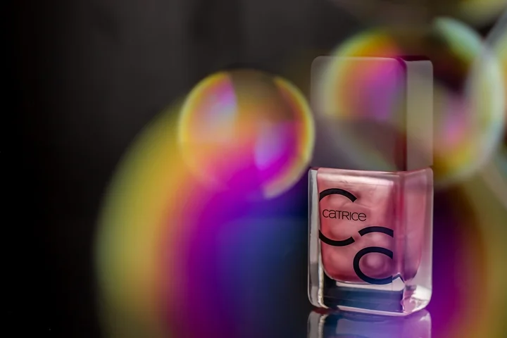 Pink nail polish bottle with a colorful, blurred bokeh background and a reflective surface.