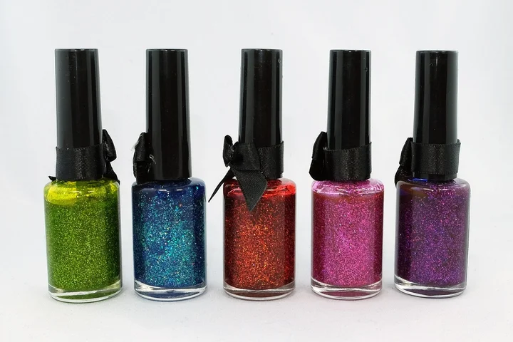 Five glittery nail polish bottles in green, blue, red, pink, and purple arranged in a row on a white background.