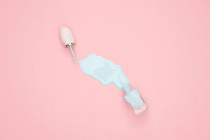 Blue nail polish spilled from an open bottle onto a pink background, with a brush nearby