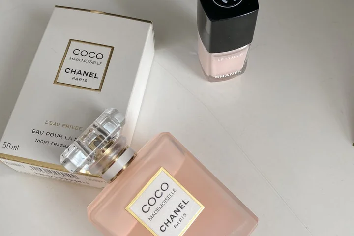 Pink nail polish bottle with Chanel Coco Mademoiselle perfume bottles in the background on a light surface.