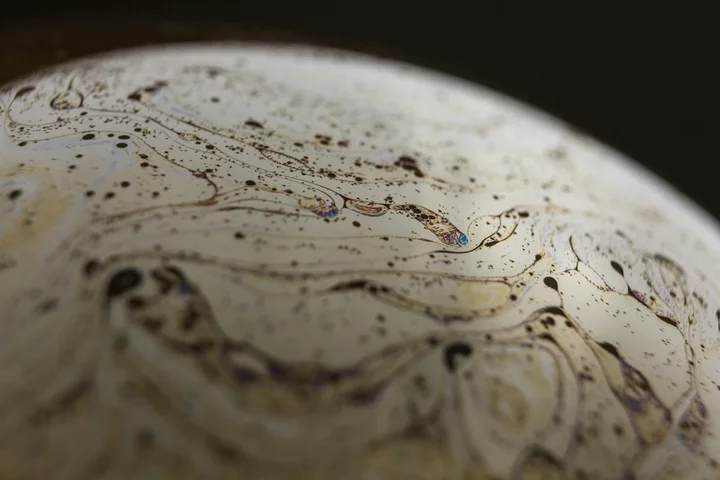 Macro close-up of oil and grease stains swirled on a light-colored surface, showing greasy patterns and droplets.