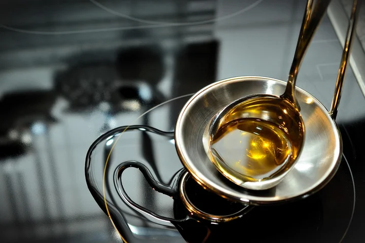 Close-up of a shiny metal dish with golden oil being poured into it on a reflective kitchen surface