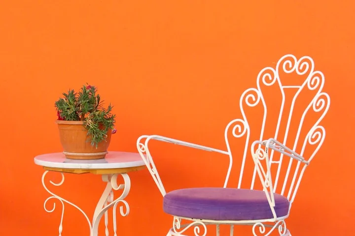 White wrought-iron patio chair with a purple cushion and a small round table with a potted plant against a bright orange wall.