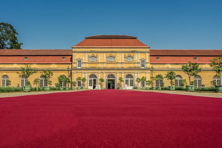 Bright red outdoor carpet spread across a courtyard in front of a yellow neoclassical building with a blue sky.