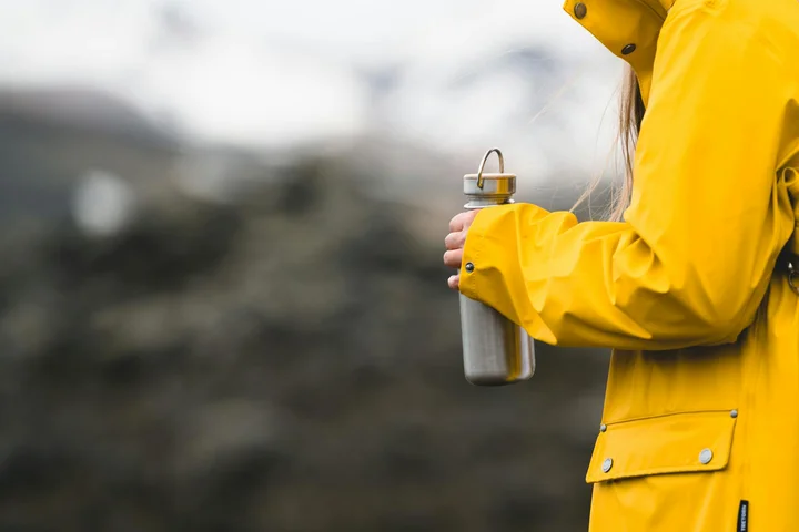 Person wearing a bright yellow rain jacket holds a metal water bottle outdoors with a blurred natural background.