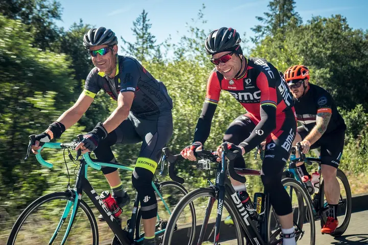 Three cyclists wearing helmets and cycling gear ride along a sunny road.