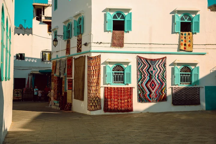 White building with turquoise window shutters and colorful rugs hanging to dry along the exterior wall