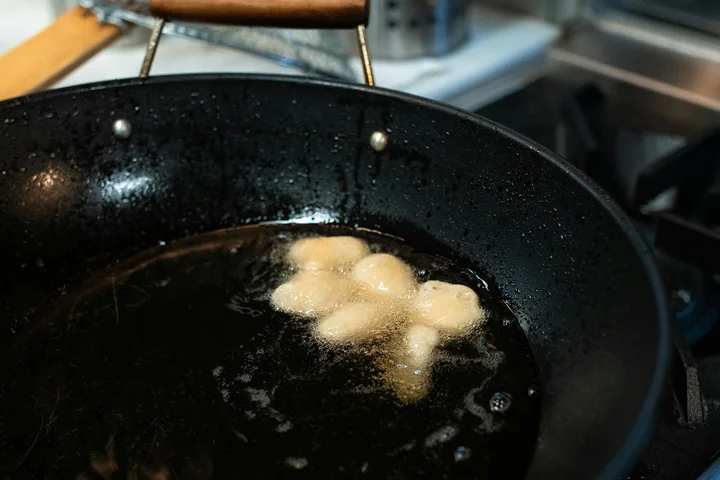 A black frying pan with hot oil and a few pale, round pieces floating on the surface; some burnt residue along the rim.