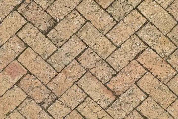 Close-up of tan brick pavers arranged in a herringbone pattern with dirt and light staining.