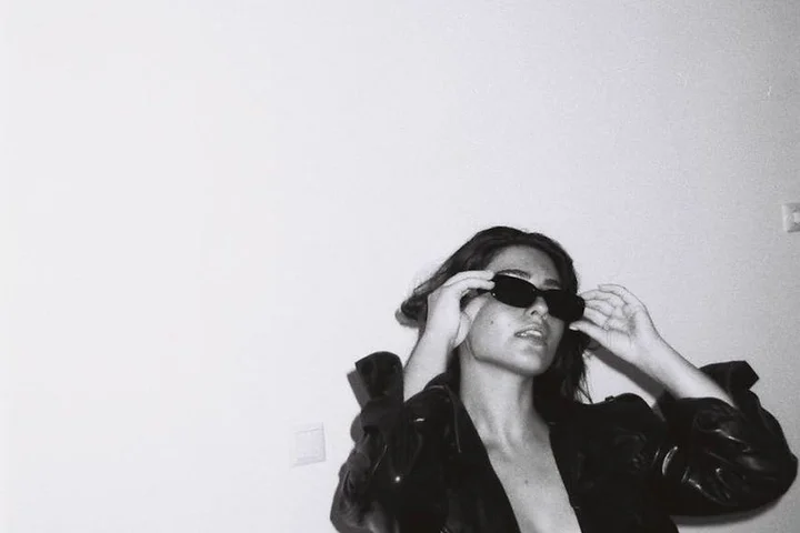 Black-and-white photo of a woman wearing sunglasses and a dark jacket, adjusting her sunglasses indoors.