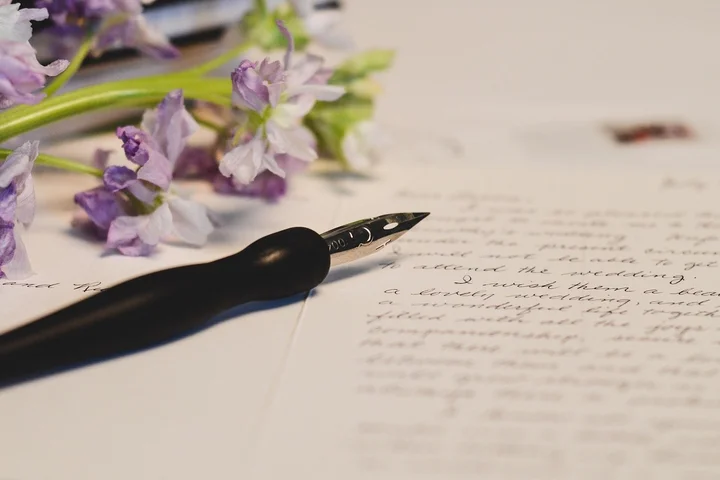 Close-up of a fountain pen resting on a sheet of handwritten paper with purple flowers in the blurred background