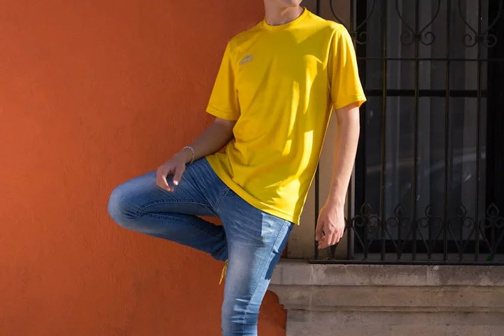 Person wearing a yellow T-shirt and blue jeans standing against an orange wall outdoors