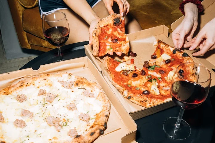 Pizza slices in open boxes on a table with two glasses of red wine and hands tearing a slice.