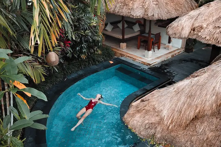 A person swimming in a curved blue pool surrounded by tropical plants and thatched-roof cabanas.