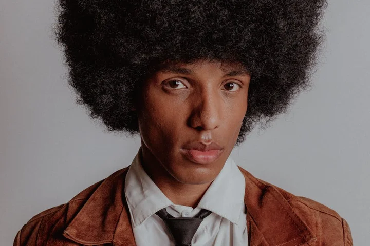 Head-and-shoulders portrait of a person with a large afro, wearing a brown jacket over a white shirt and black tie, against a neutral gray background.