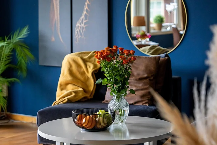 Living room after renovation: blue accent wall, round mirror, navy sofa with a yellow throw, white round coffee table with a vase of orange flowers and a bowl of fruit.