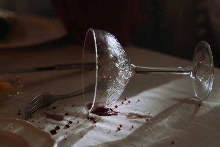 A wine glass tipped over on its side with red wine spilled on a white tablecloth, and a few droplets nearby.