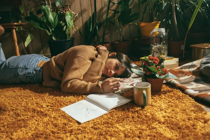 A person lying on a bright orange shag carpet indoors with papers, a mug, and plants around.
