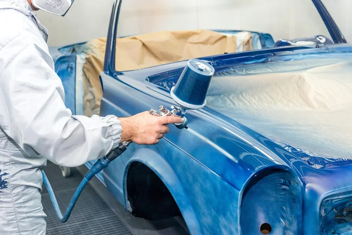 Technician in protective gear uses a spray tool to clean tar from a blue car's painted surface inside a workshop.