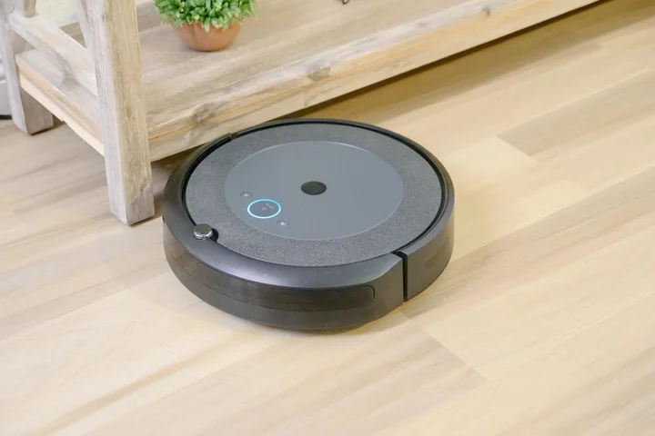 Black circular robot vacuum on a light wood floor near a wooden shelf.