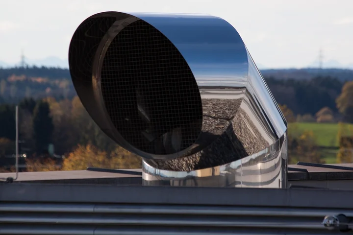 A curved metal turbine roof vent on a house with a clear blue sky and autumn trees in the background.