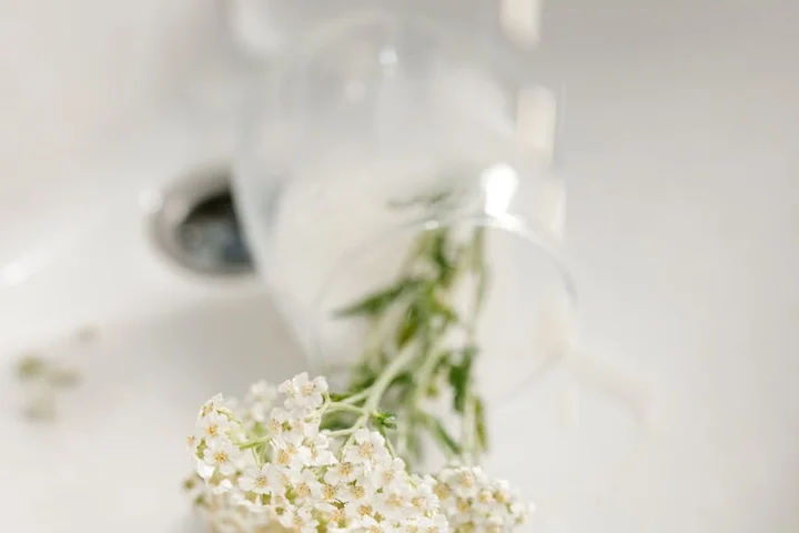 Close-up of a clear spray bottle with a blurred white sink in the background and a small bouquet of white flowers in the foreground.
