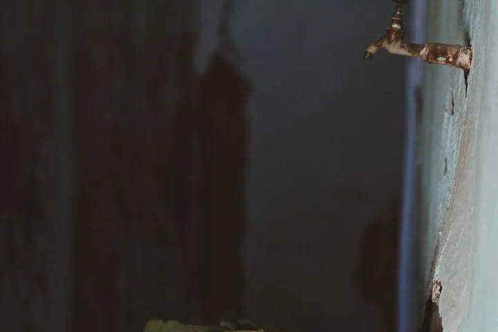 Dim indoor scene with a rusty metal pipe protruding from a wall and a dark, shadowy background.