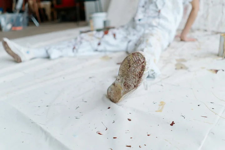 Close-up of a paint-splattered sneaker on a white surface with a person wearing paint-splattered clothing in the background.