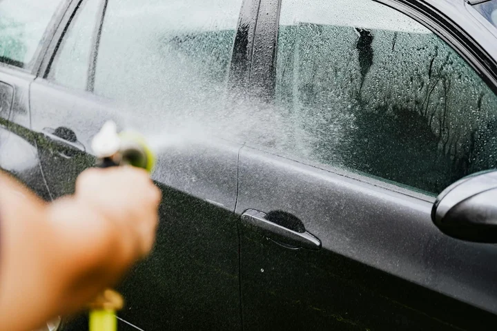 A hand holding a spray bottle aimed at a car door, spraying liquid with water droplets on the window.