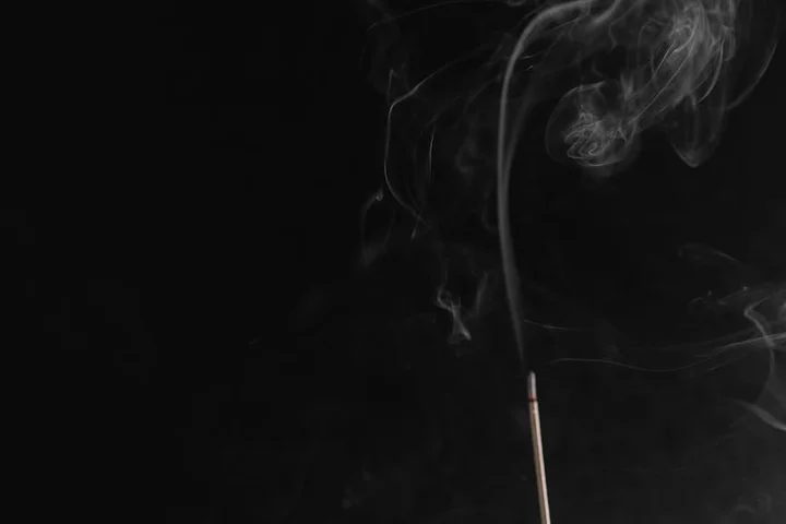 Close-up of a lit incense stick releasing curling wisps of smoke against a dark background.