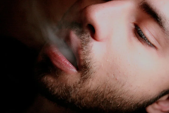 Close-up of a bearded person exhaling cigarette smoke.