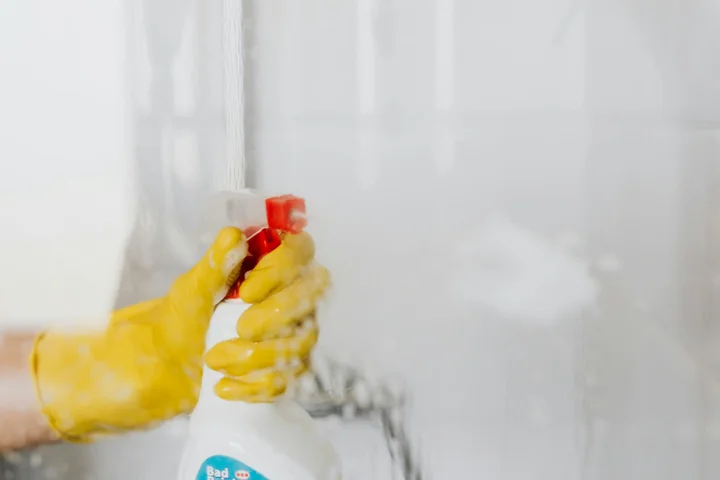 Hands wearing yellow cleaning gloves gripping a spray bottle and releasing cleaner onto a foggy glass surface