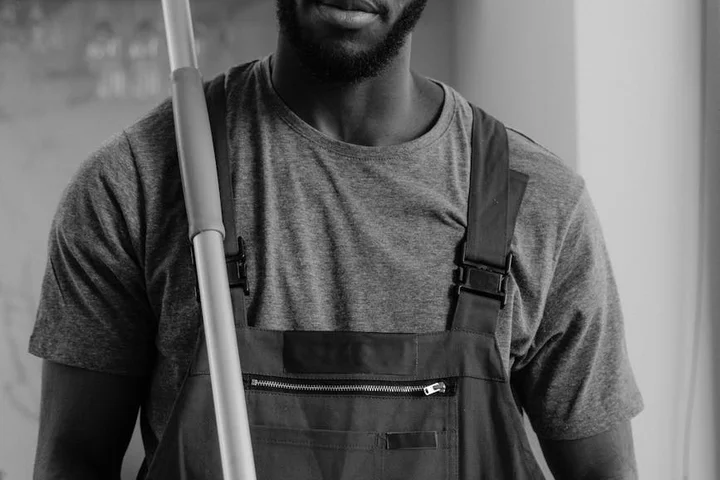 Black-and-white image of a person wearing work overalls holding a mop handle