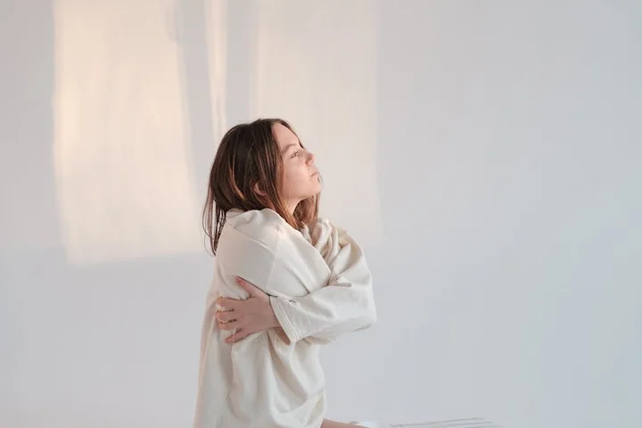 Person standing in a softly lit room with arms crossed over the chest, wearing a light sweater, looking up with a calm expression.