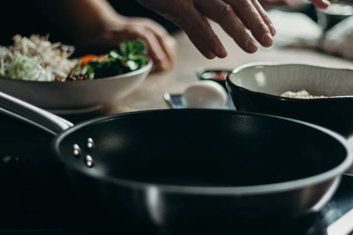 Close-up of a frying pan on a stove top with hands preparing ingredients in the background.