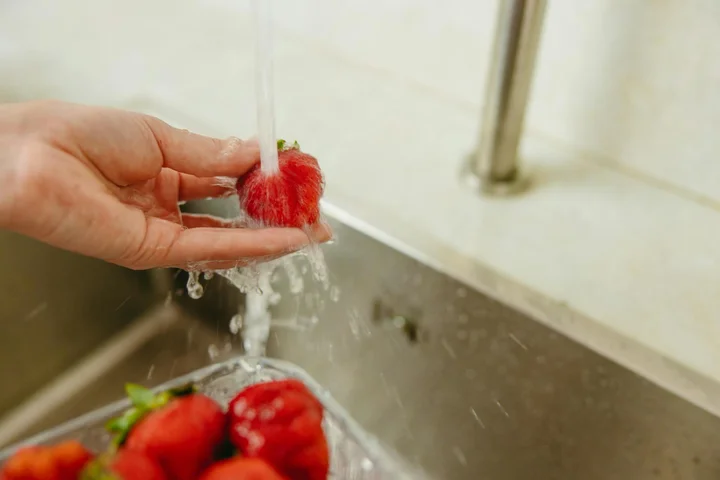 Hand holding a fresh strawberry under a stream of running water in a kitchen sink.