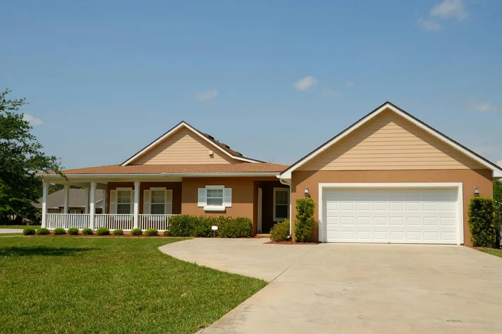 Suburban single-story house with a curved concrete driveway leading to a two-car garage, beige siding, and a well-kept lawn under a clear blue sky.