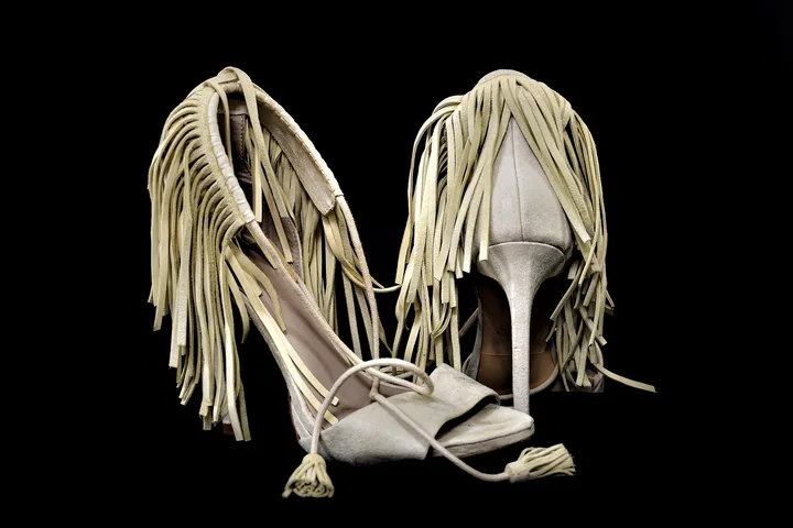 Beige suede fringe high-heeled sandals positioned against a black background.