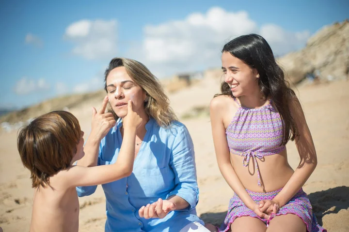 Two adults and a child on a sunny beach; a child applies sunscreen to one adult while the other adult sits nearby.