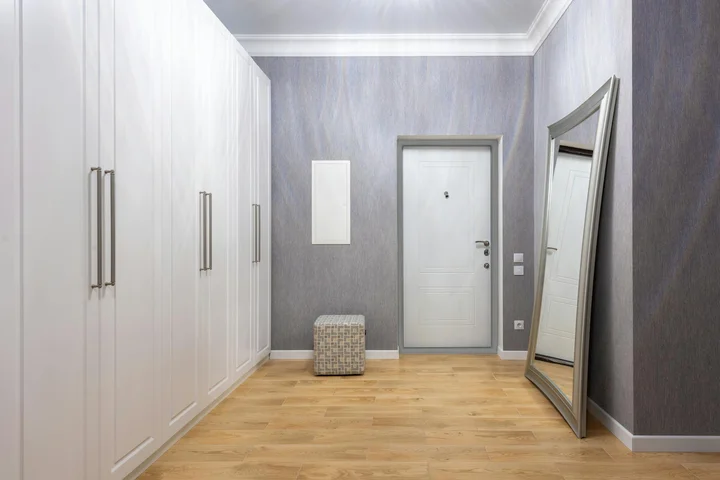 Interior of a bright closet or hallway featuring white built-in wardrobes on the left, a light-colored wooden floor, a standing mirror on the right, and a small ottoman near the door at the end.