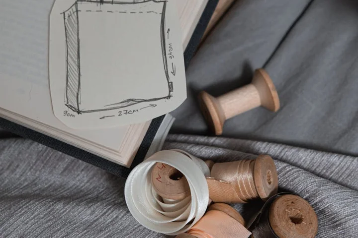 Sketch of fabric dimensions with an open notebook and scattered vintage thread spools, illustrating materials involved in care testing for textiles.