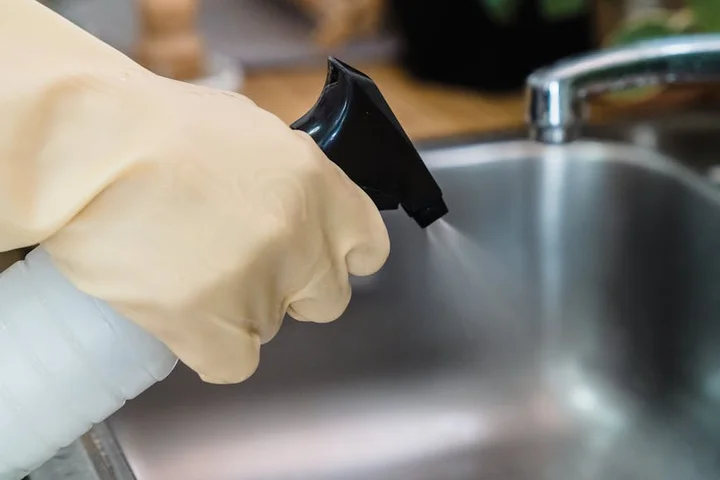 Gloved hand spraying cleaner at a stainless steel kitchen sink.