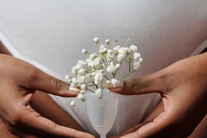 Close-up of hands forming a frame around a small bouquet of white baby's breath set inside a white menstrual cup against a light background.