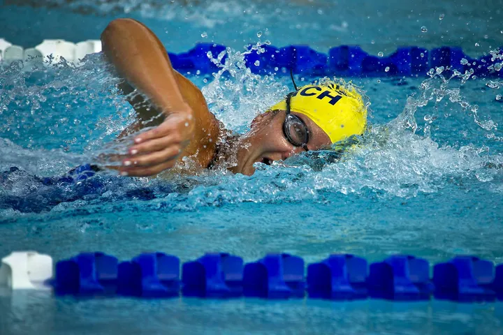Swimmer wearing a yellow cap and goggles gliding through pool water with blue lane markers in the background.