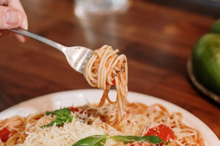 A hand holds a fork twirling spaghetti with tomato sauce on a plate, sprinkled with grated cheese and basil.