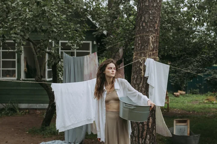 A woman stands outdoors beside a clothesline hung between trees, holding a gray bucket with white fabrics drying on the line behind her.